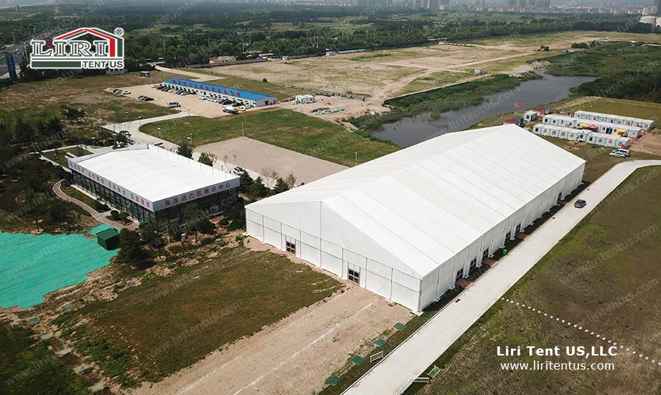 Aerial view of a large white A-frame clearspan structure on a construction site, surrounded by open land and temporary buildings, showcasing the durability and scale of Liri Tent US structures.